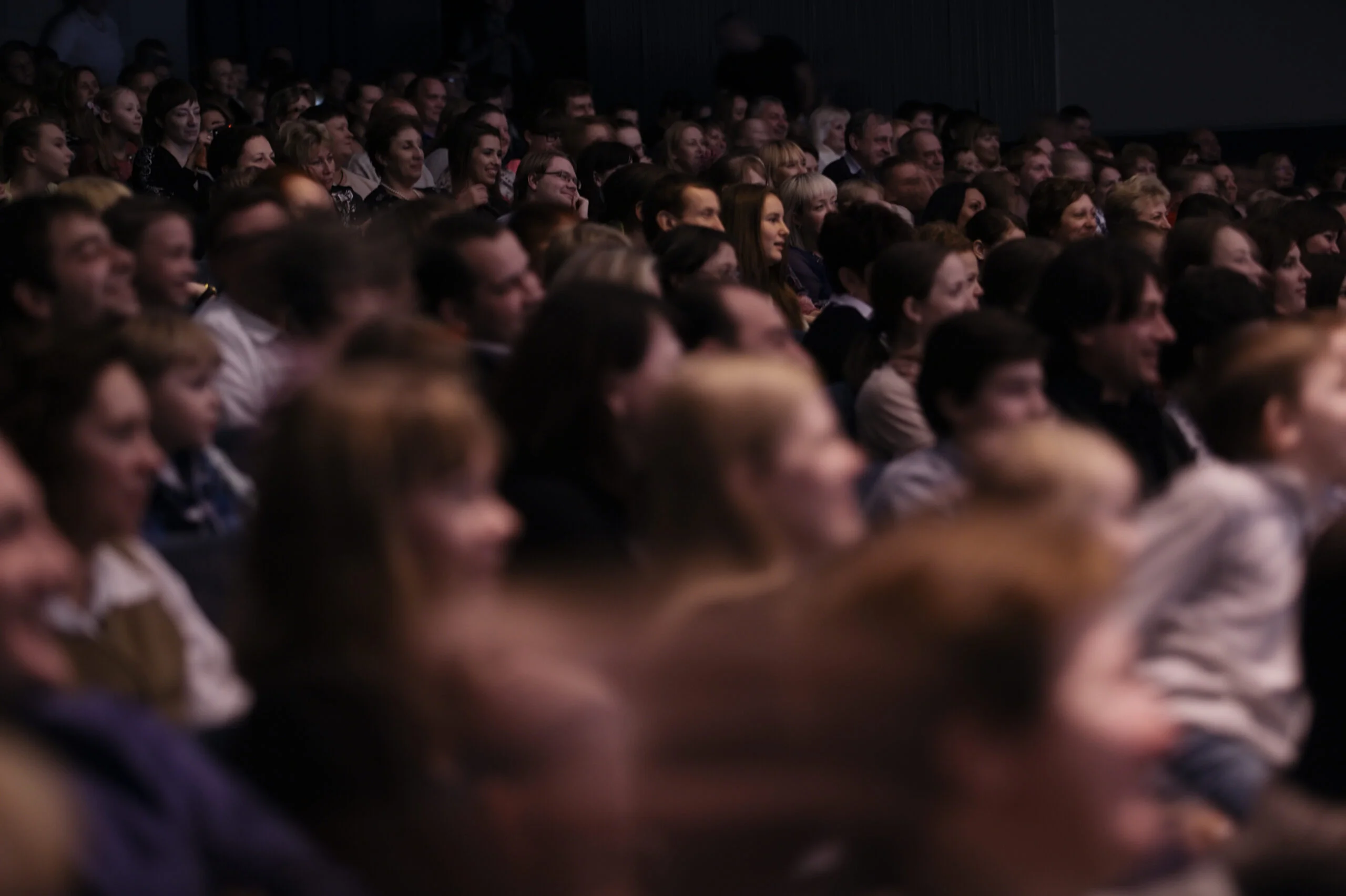 A large audience seated in a theater, attentively watching a performance or presentation. The crowd is dimly lit, creating a focused and immersive atmosphere.