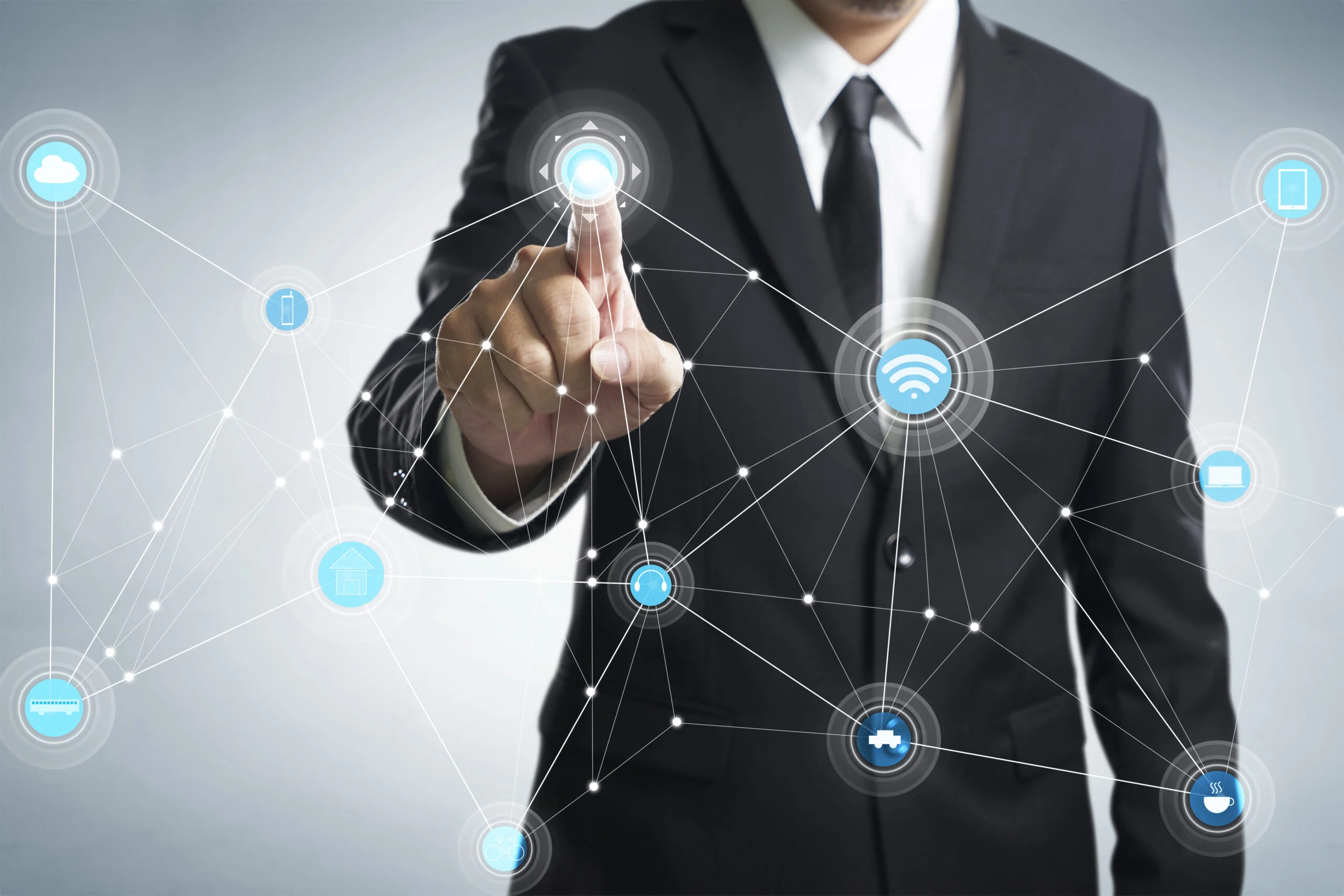 A man in a suit points to a digital network with connected icons representing various technologies, such as Wi-Fi and smart devices, on a transparent interface. The background is a gradient of gray and white.