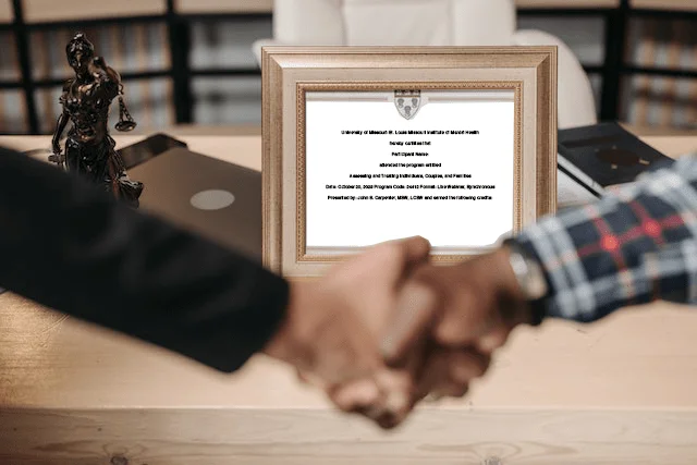 Two people are shaking hands over a desk. In the background, a framed certificate and a standing statue are visible, along with some books and folders. The setting appears to be an office or study.