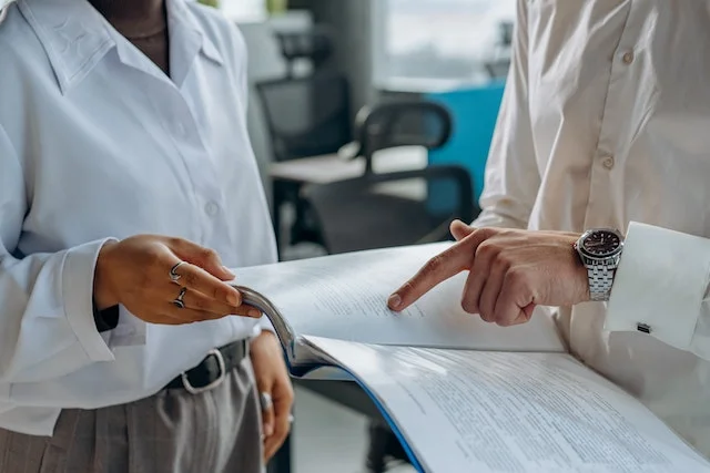 Two people in professional attire stand close, looking at a document. One person points at the page, wearing a watch. The background shows an office setting with chairs and desks.